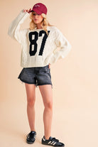 Aemi+Co Contrast Number Cable Knit Drop Shoulder Sweater featuring a sporty 87 design, paired with denim shorts and a cap.