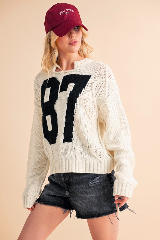 Aemi+Co Contrast Number Cable Knit Drop Shoulder Sweater with bold 87 design, worn with a casual cap and shorts.