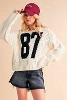 Aemi+Co Contrast Number Cable Knit Drop Shoulder Sweater in cream with black number 87, styled with a maroon cap and denim shorts.