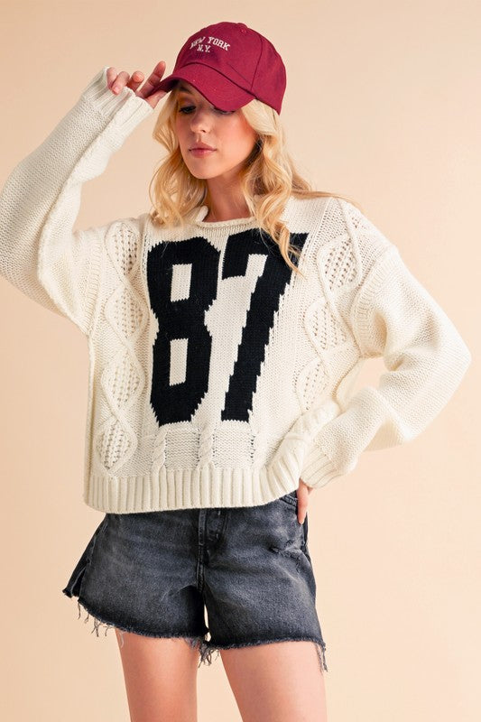 Aemi+Co Contrast Number Cable Knit Drop Shoulder Sweater in cream with black number 87, styled with a maroon cap and denim shorts.