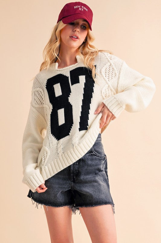 Aemi+Co contrast number cable knit drop shoulder sweater in cream with bold 87, styled with a black denim skirt and red cap.