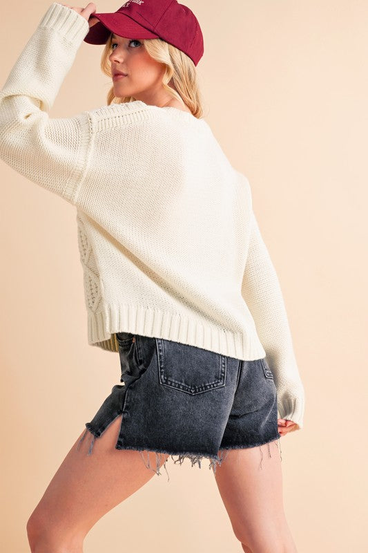 Model showcasing Aemi+Co Contrast Number Cable Knit Drop Shoulder Sweater paired with stylish denim shorts and a cap.